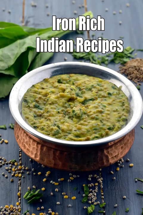 A traditional copper serving bowl filled with a vibrant green spinach and lentil dal sits on a dark blue wooden surface. The dish appears thick and hearty, containing visible bits of chopped spinach and small pulses. Scattered around the base of the bowl are raw lentils, seeds, and fresh green herbs. In the background, a bundle of fresh leafy spinach and a small pile of cumin seeds are visible, emphasizing the healthy ingredients. Large white text at the top reads, Iron Rich Indian Recipes.