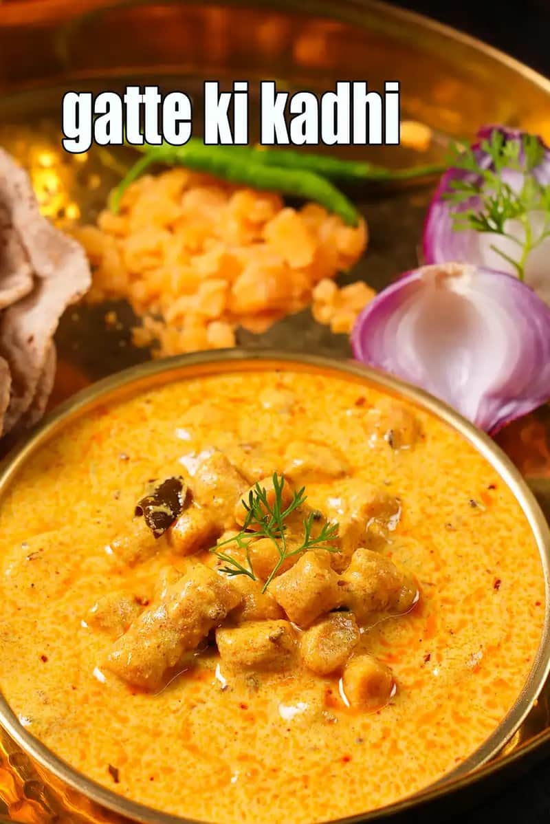 A bowl of traditional Rajasthani Gatte ki Kadhi served in a brass bowl, showing gram flour dumplings simmered in a rich yellow yogurt-based curry, garnished with herbs, with chapati, green chilies, onion, and condiments placed around it.