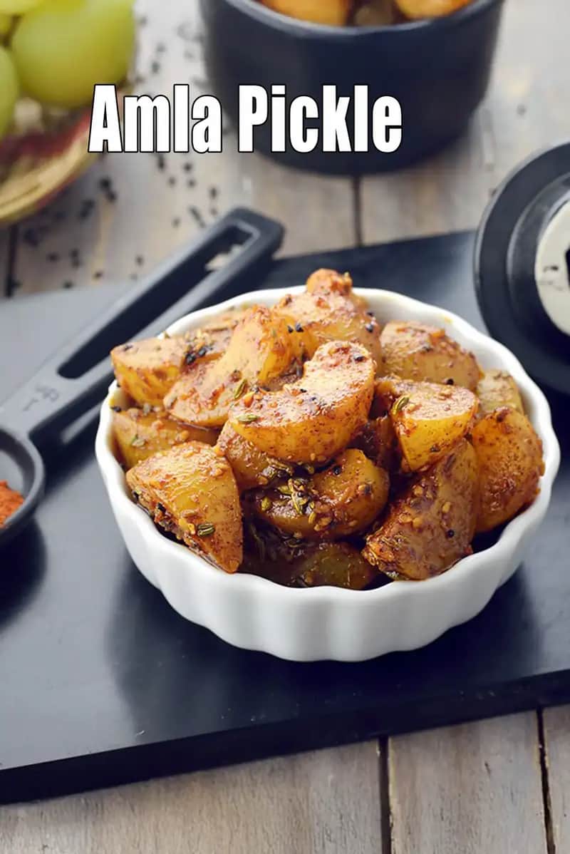 The image shows a close-up of a white bowl filled with segmented Amla pickle, coated in a rich, reddish-brown spice mix including fennel seeds and kalonji. In the background, there are fresh green amlas in a basket and a black kitchen utensil, all set on a dark, rustic surface.