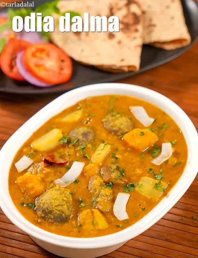 An image featuring a white oval bowl filled with Odia Dalma, a traditional lentil and vegetable stew from Odisha. The thick, mustard-colored gravy is packed with chunky pieces of cooked vegetables, including potatoes and pumpkins, and is garnished with fresh green coriander and thin slivers of fresh coconut. In the background, a plate with soft rotis, fresh tomato slices, and onion rings is visible, resting on a rustic wooden surface.