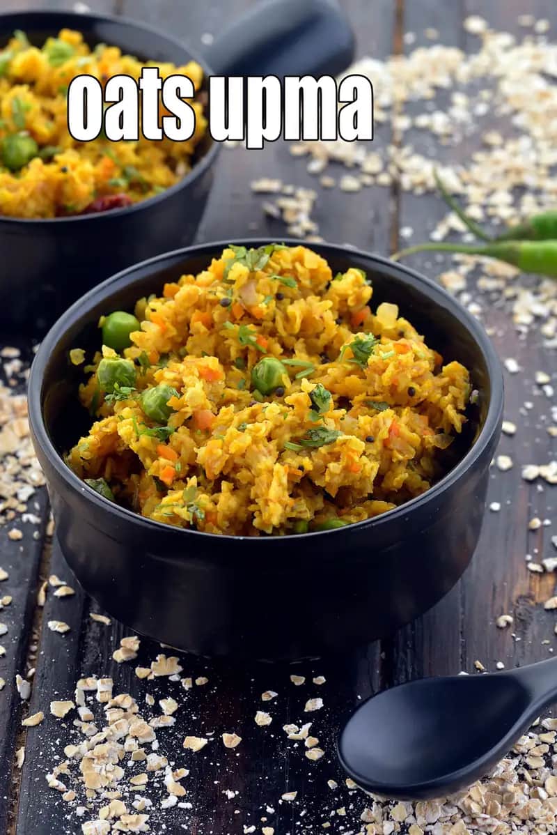 A close-up shot of a black bowl filled with a savory and colorful oats upma, garnished with fresh green peas and coriander. In the background, another bowl of the same dish is partially visible, along with scattered raw rolled oats and a green chilli on a dark, rustic wooden surface.
