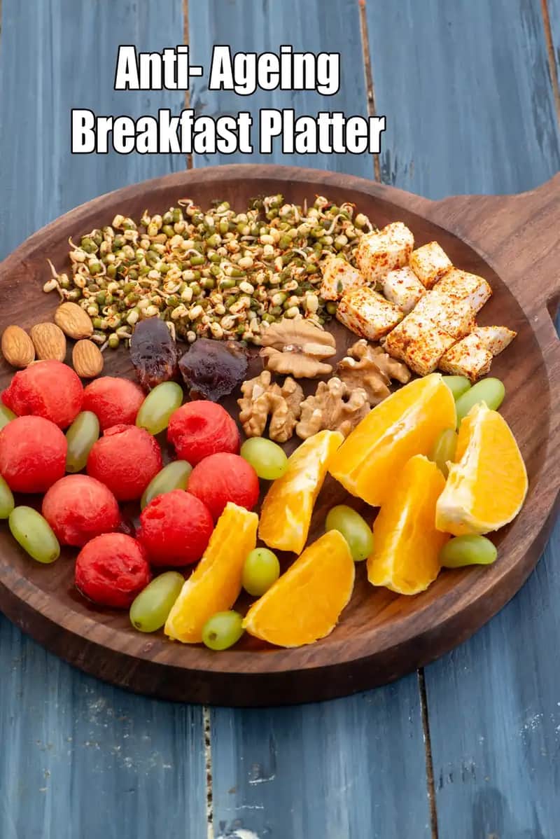 An image of an Anti-Aging Breakfast Platter served on a round dark wooden board with a handle, placed on a blue rustic wooden surface. The platter is neatly arranged with various healthy components: vibrant orange wedges, green grapes, and watermelon scoops on one side. The other half features protein and fiber-rich items, including a pile of sprouted moong beans, spiced paneer cubes, whole walnuts, almonds, and dates.