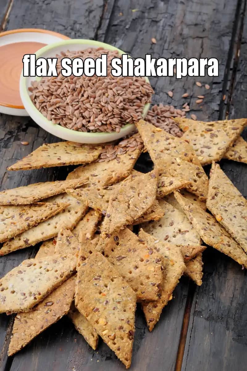 An image of Flax Seed Shakarpara, featuring a heap of diamond-shaped, baked savory crackers speckled with flax seeds, scattered across a dark, rustic burnt-wood surface. In the background, there is a small white bowl filled with raw brown flax seeds, with a few seeds spilled around it.