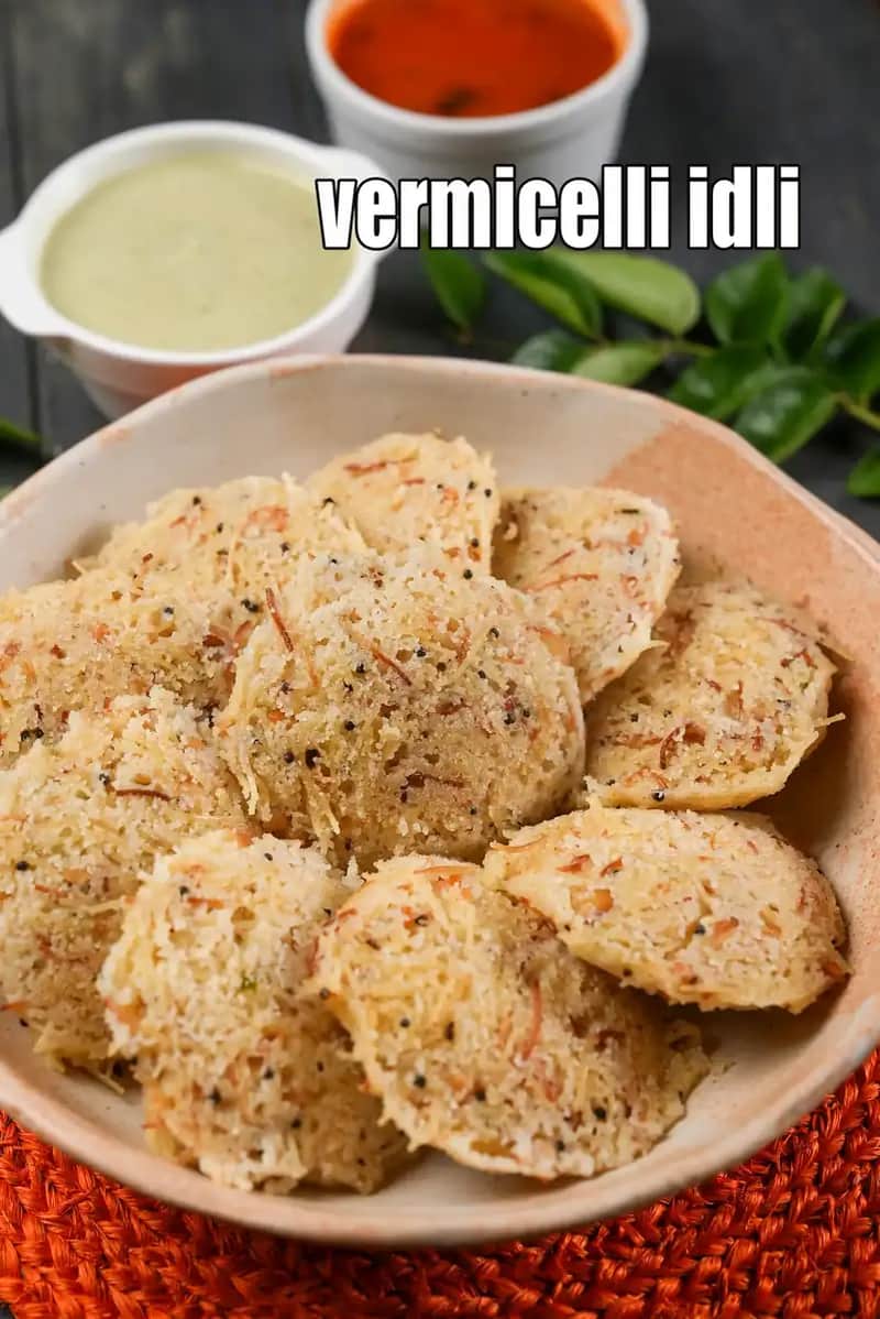 A bowl of soft vermicelli idli made from roasted semiya, garnished with mustard seeds and spices, served with coconut chutney and red chutney in the background on a dark surface.