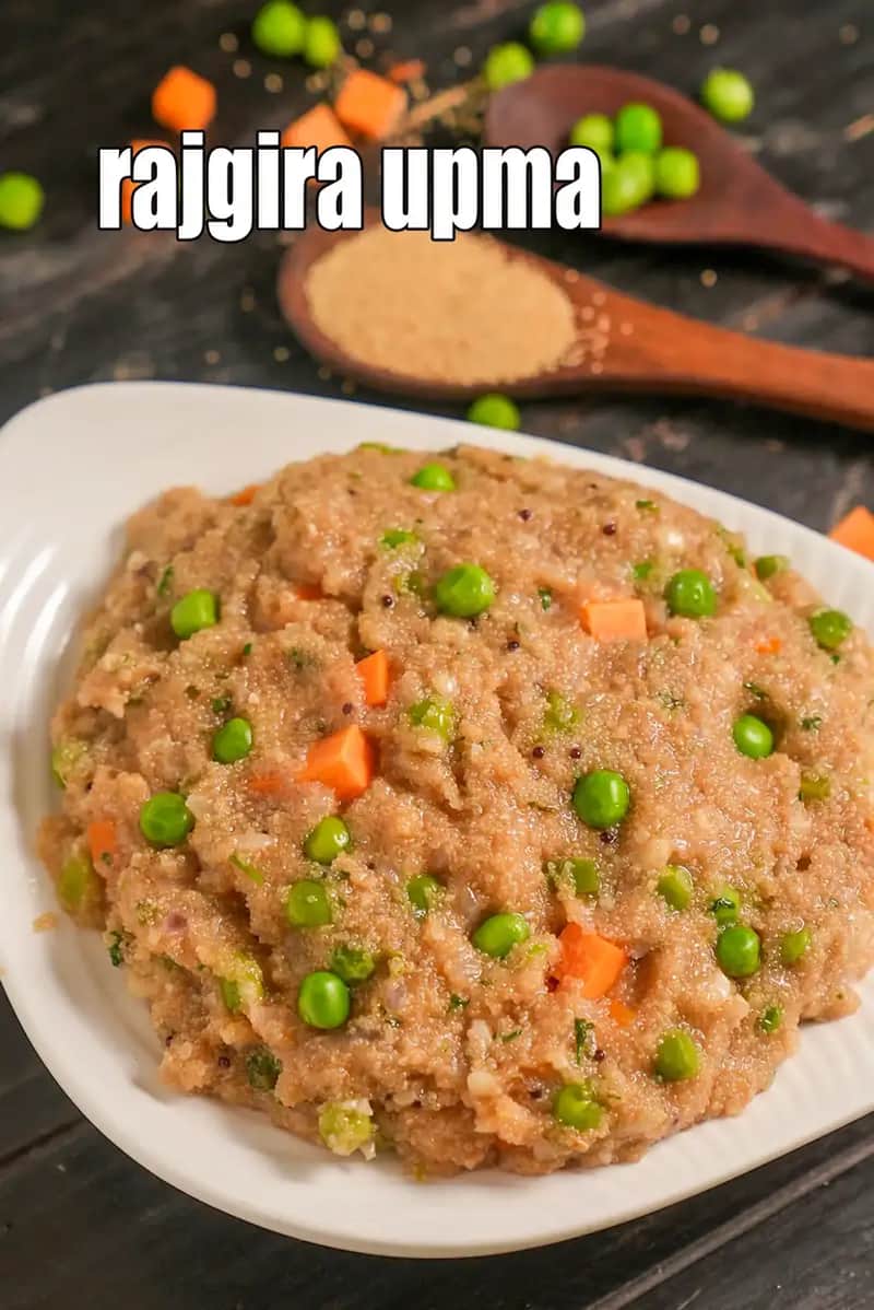 An image showing a close-up of Rajgira Upma (Amaranth Upma) served on a white plate. The dish has a grainy, light-brown texture and is mixed with bright green peas and diced orange carrots. In the background, wooden spoons hold more peas and raw rajgira grains, scattered across a dark, rustic surface.
