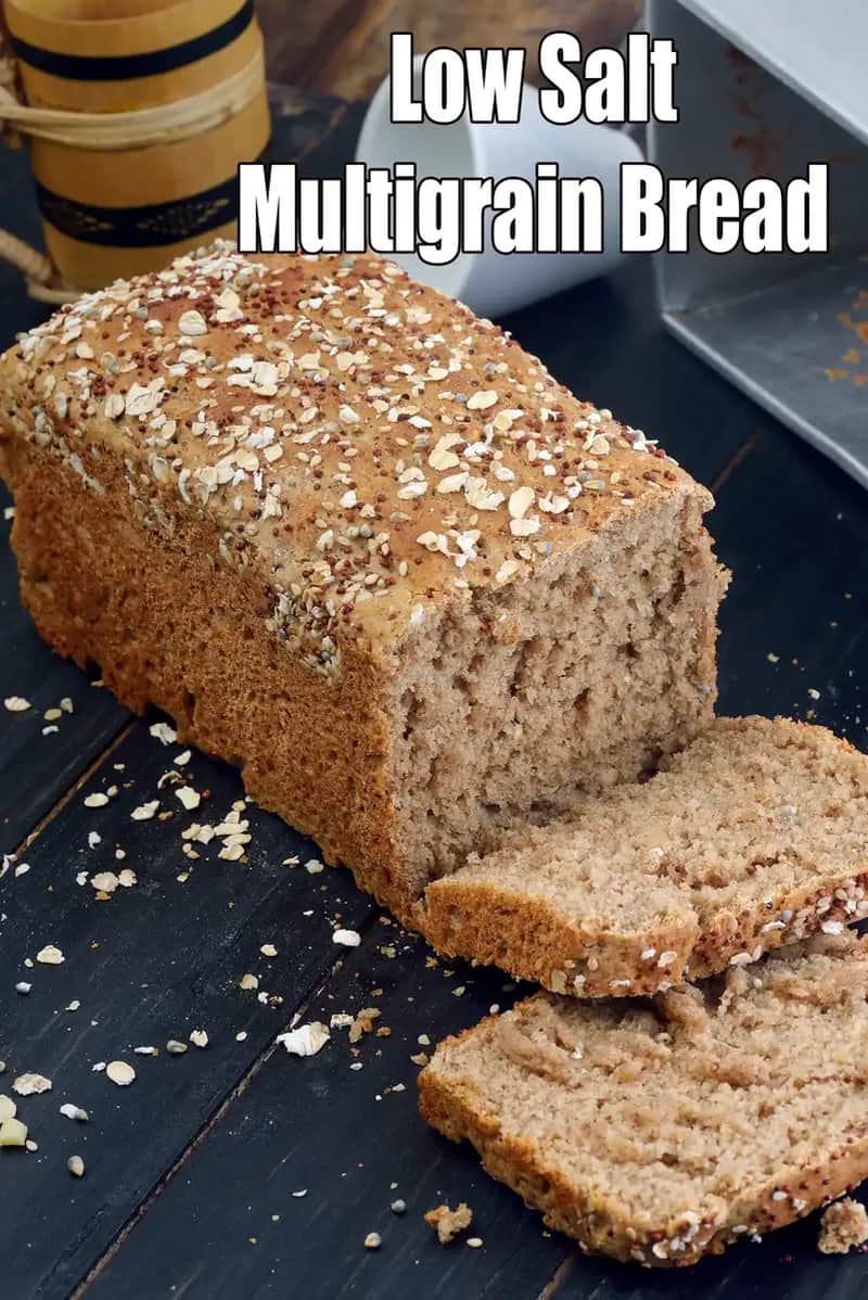 A rectangular loaf of low-salt multigrain bread sits on a dark, rustic wooden surface. The bread has a thick, hearty golden-brown crust topped with a generous layer of oats, seeds, and grains.