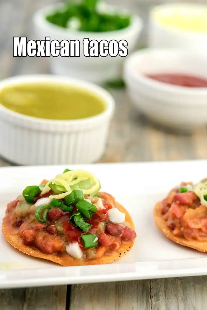 A close-up shot of two crispy, open-faced Mexican Tacos (tostada-style) served on a white plate. The tacos are topped with a savory red bean filling, chopped white onions, fresh green scallions, and a drizzle of red salsa. In the background, small white bowls filled with green dip and red sauce are visible, creating a colorful and appetizing spread.
