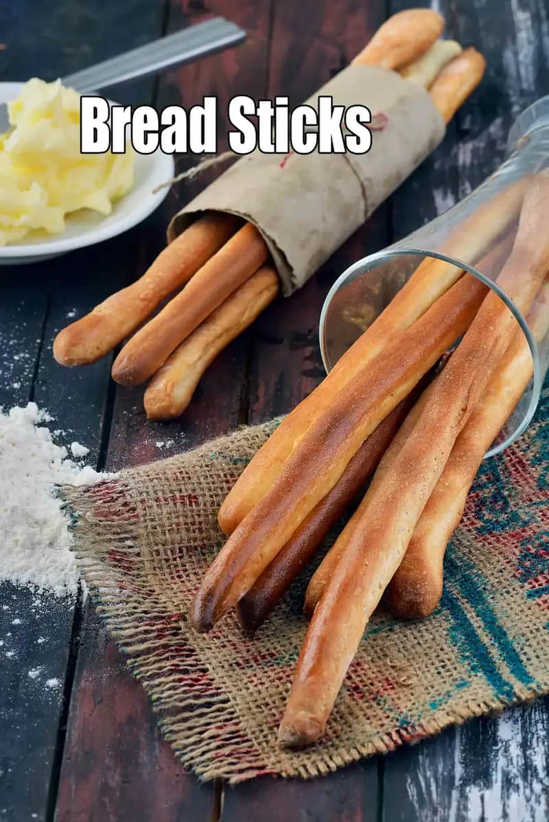 A close-up shot of golden-brown, crispy homemade Indian breadsticks (Grissini) stacked neatly on a wooden platter. The breadsticks are speckled with cumin seeds (jeera) and dried herbs. In the background, there is a small ceramic bowl of creamy cheesy dip or spicy chutney, with a warm, rustic kitchen setting that emphasizes their crunchy texture.