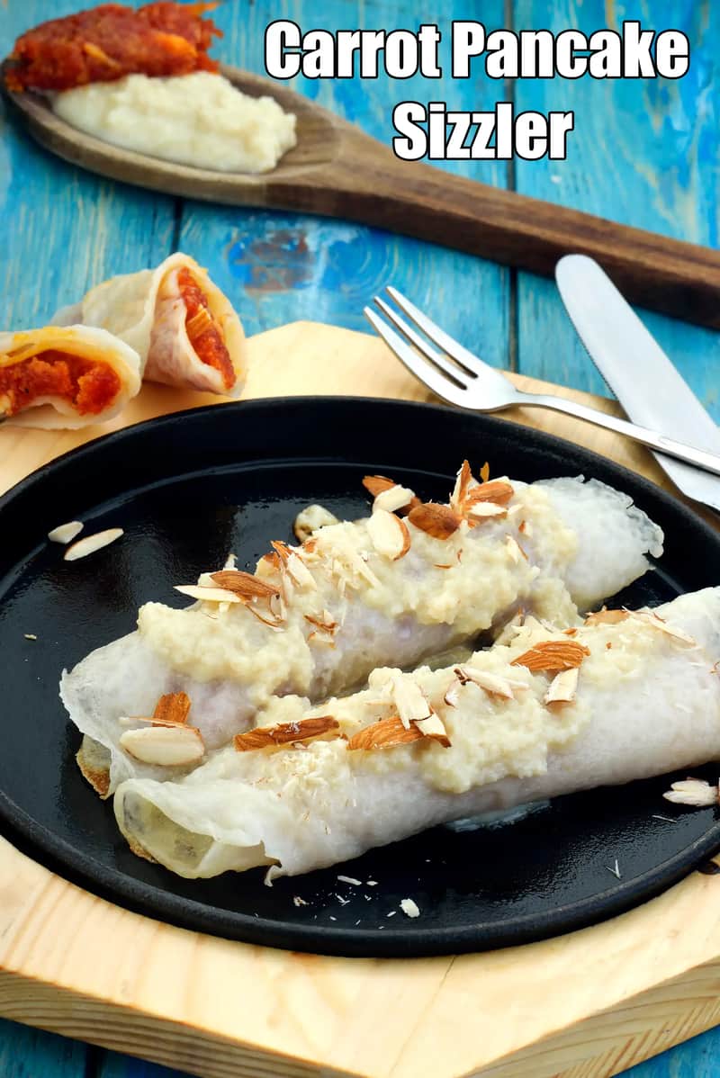 Carrot Pancake Sizzler served on a black sizzling plate, topped with creamy white sauce and garnished with sliced almonds, with carrot-stuffed rolls and cutlery placed on a blue wooden background.