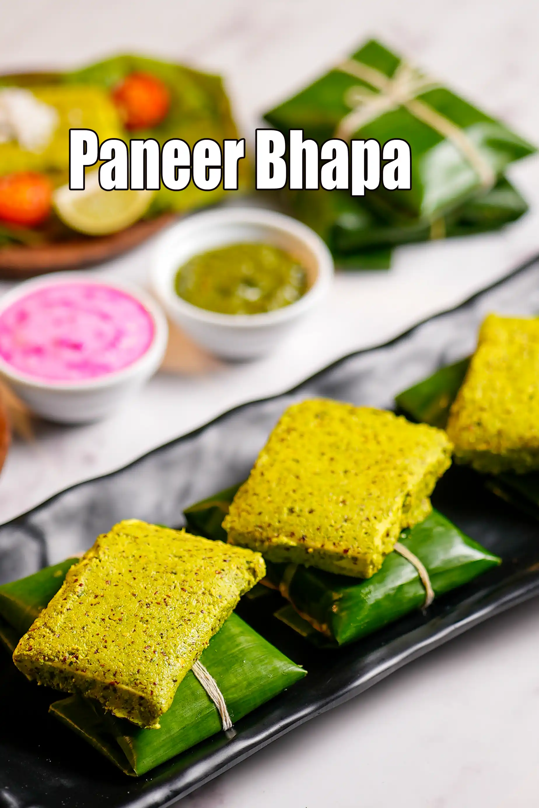 Paneer Bhapa served on a black platter, showing steamed paneer pieces wrapped in banana leaves, coated with green mustard paste, accompanied by green chutney and pickled onions.