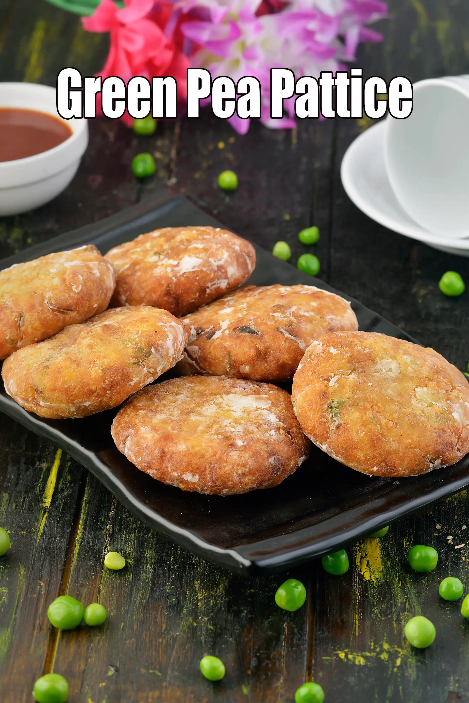 A black square plate holding golden-brown green pea pattices arranged neatly, placed on a dark wooden surface. Fresh green peas are scattered around the plate, with a small bowl of chutney and white cups in the background, giving a rustic food presentation.