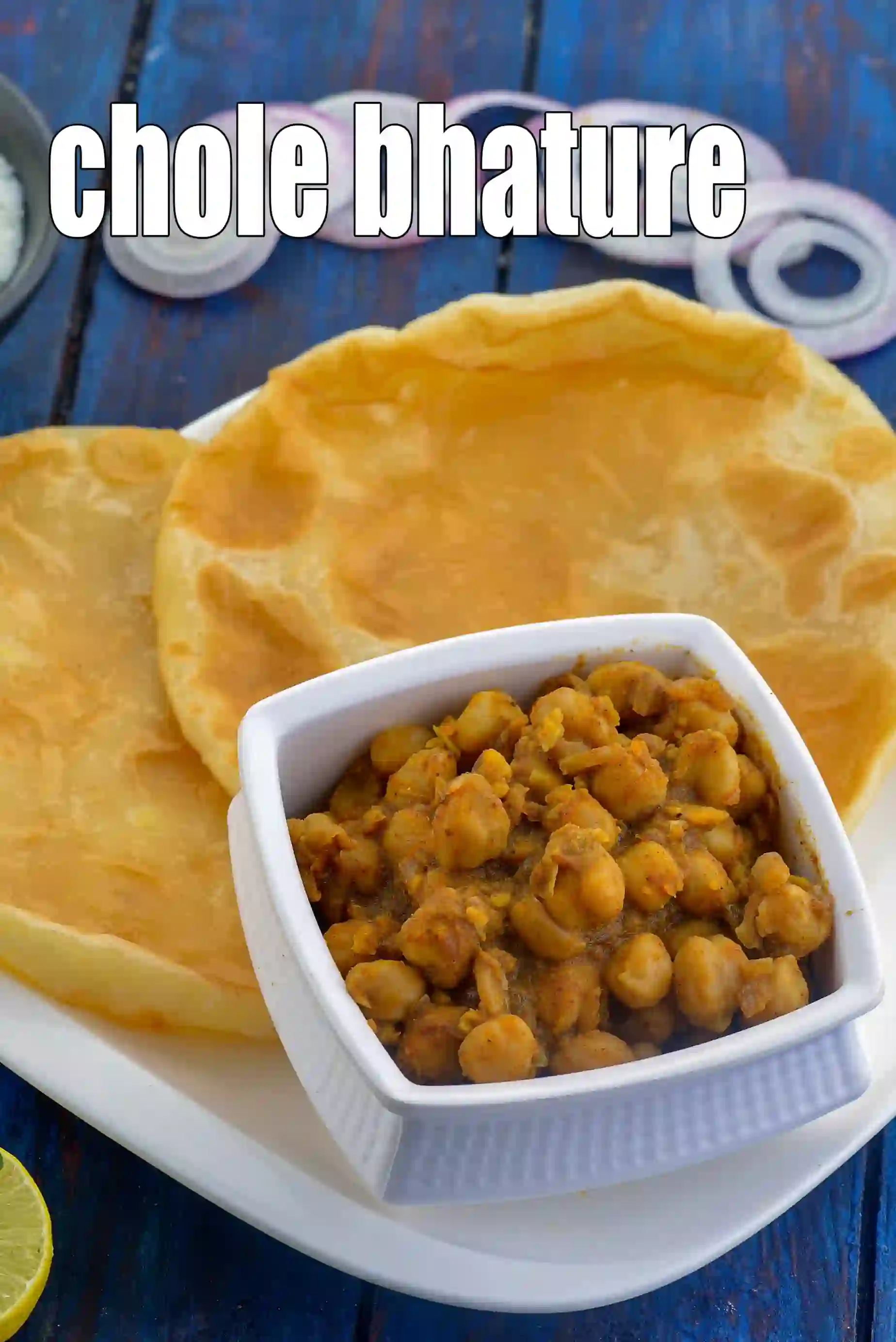 A plate of traditional Chole Bhature served with fluffy, golden-fried bhature and a bowl of spicy chickpea curry (chole), garnished with onion slices and lemon on a rustic blue wooden background.