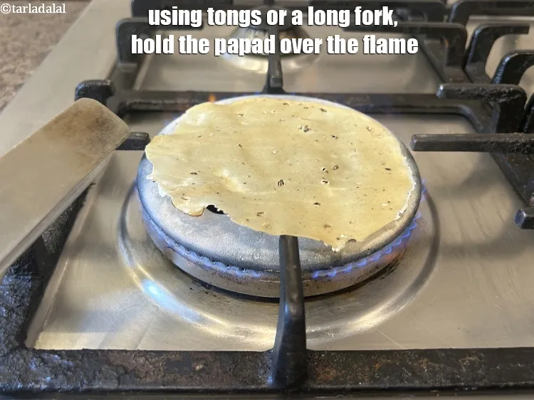 Step 10 – <meta charset="UTF-8" />Using tongs or a long fork,&nbsp;hold the papad over the flame.