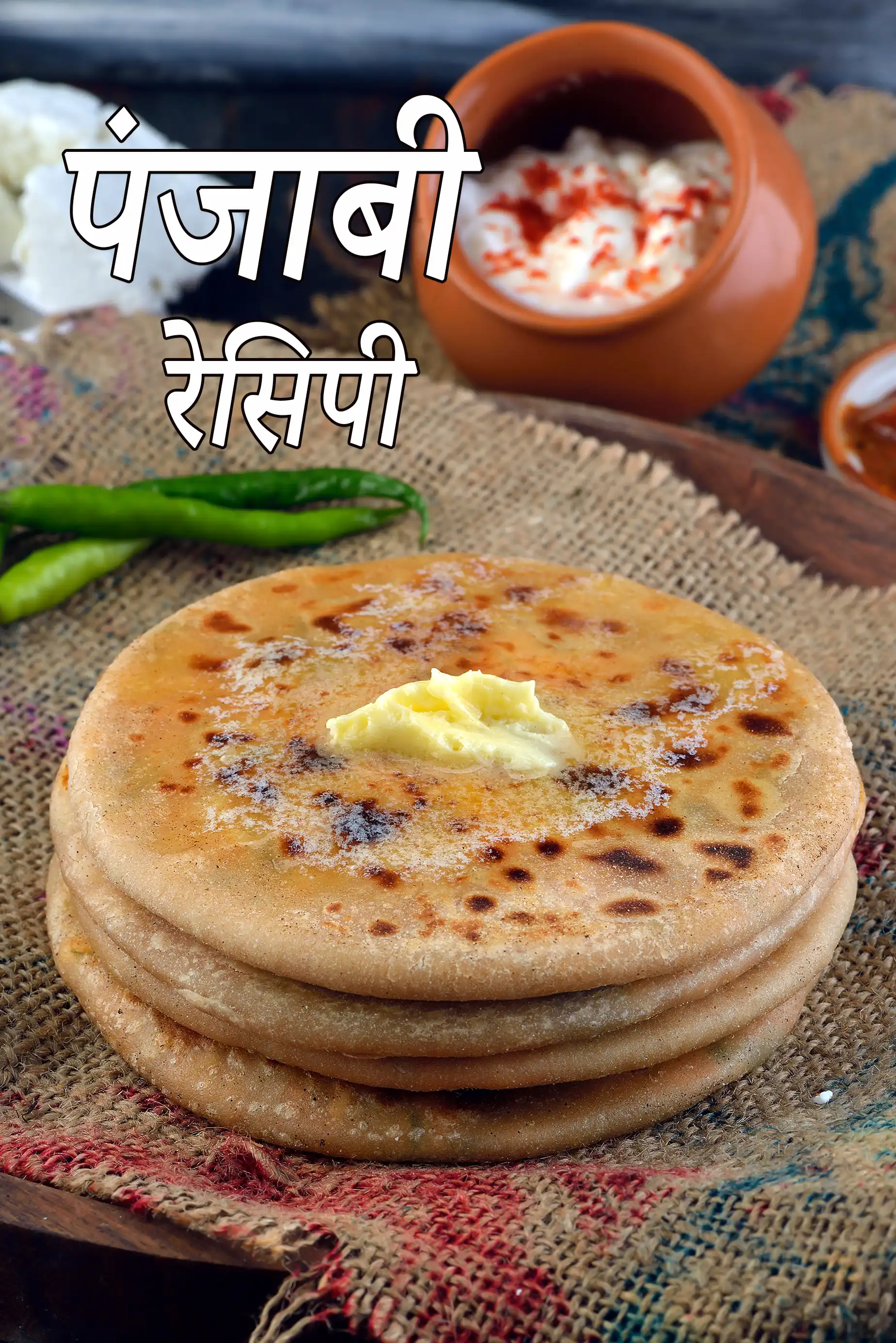 मक्खन से सजी हुई ताज़ी बनी हुई भारतीय स्टफ्ड पराठों की एक परतदार ढेरी, जिसे देसी कपड़े पर परोसा गया है, साथ में हरी मिर्च, दही और अचार रखा है।